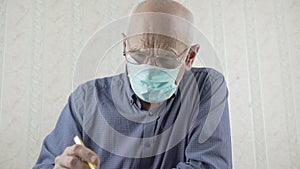 Gray haired old man with glasses sits at table and writes letter with pen