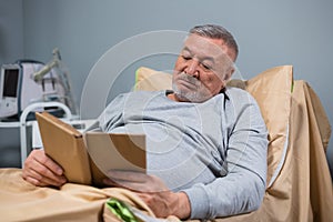 A gray-haired old man reading a book in his bed in a hospital ward