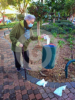 A gray-haired old woman with a stick cleans up the thrown away papers into a street bin