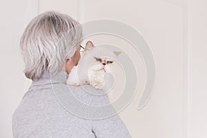 A gray-haired older woman in a gray turtleneck has a cute beige cat on her shoulder. The view from the back.