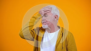 Gray-haired pensioner showing face-palm gesture against orange background, fail