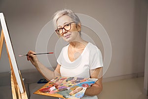 Gray-haired sophisticated lady in glasses stands in front of easel