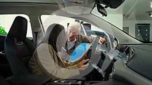 A gray-haired woman gets behind the wheel. An elderly Caucasian couple chooses a new car at a car dealership.