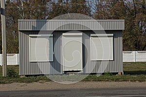 Gray metal building with windows and a door closed by white roller blinds on green grass