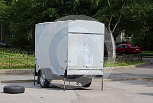 A gray metal closed two-wheeled trailer for a passenger car is standing on the street