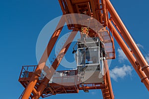 Gray mobile cab with lifting hook, located on rail of huge orange overhead crane, electric shields are mounted on left.