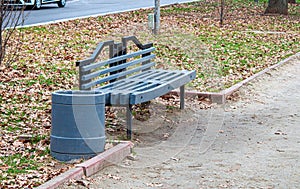 A gray painted wooden bench and a trash can in an autumn park near the footpath