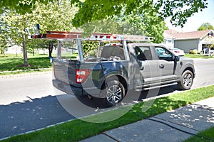 Gray pickup truck with ladders on a rack and equipment in the bed parked by a road curb