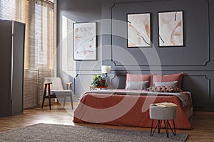 Gray, retro armchair standing in the corner of an elegant bedroom interior with watercolor posters on dark gray wall with molding