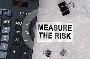 On a gray surface lies a calculator, a pen and a business card with the inscription - Measure The Risk