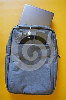 Gray textile city backpack containing silver laptop, smartphone and an external battery - power bank on a yellow background. Study