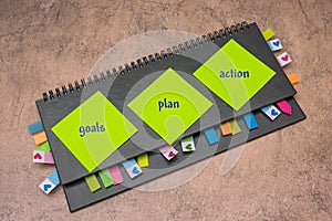 Gray weekly planner with bookmarks and stickers on a brown background. On yellow stickers it is written - goals, plan, action.