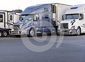 Gray and white big rig semi trucks with semi trailers standing in row on truck stop parking lot