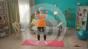 Grayhaired elderly woman doing fitness with dumbbells at home. A mature woman build up biceps, trains triceps, keeps her