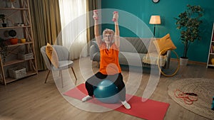 A grayhaired retired woman sitting on a fitness ball performs an exercise with dumbbells. An elderly woman raises