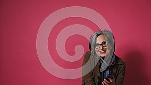 Graying pretty woman take phone down smiles. Smart mature lady in eyeglasses and brown jacket on pink background. Text