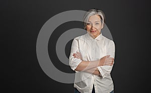 Graying pretty woman in white shirt stands arms-crossed isolated on dark gray background. High quality photo