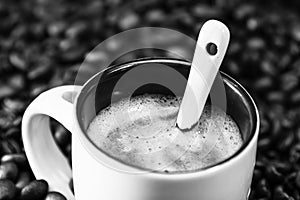 Grayscale closeup of a mug with hot expresso on roasted coffee beans