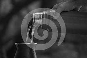 Grayscale selective focus shot of a hand pouring water from a thermos in a  calabash mate cup with straw