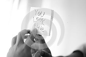 Grayscale shot of a person's hand holding a blank paper with black lettering ''You Look Fine''