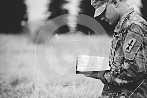 Grayscale shot of a young soldier reading a bible in a field