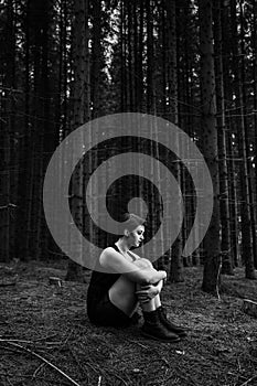 Grayscale of a young female in a black dress sitting on the ground alone in the forest