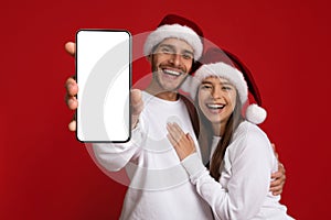 Great App. Happy Young Couple Wearing Santa Hats Showing Blank Smartphone Screen