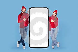 Great App. Joyful Couple Wearing Knitted Hats Standing Near Big Blank Smartphone