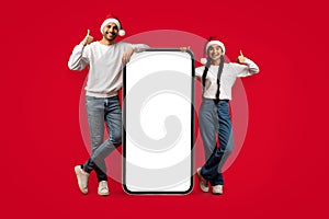 Great App. Joyful Couple Wearing Santa Hats Standing Near Big Blank Smartphone