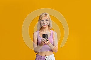 Great app. Positive young woman holding and using cellphone, chatting online isolated on yellow studio background