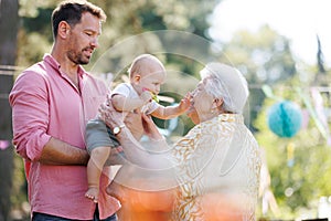 Great-grandmother holding little baby in her arms. Family summer garden party.