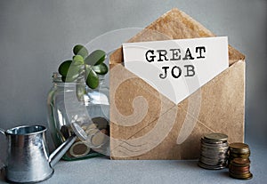 GREAT JOB text is written on white paper on an antique envelope, which lies on the table along with a stack of coins, a glass jar