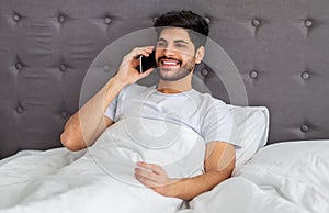 Great morning news. Happy arab man talking on smartphone with friend or lover, sitting in bed, copy space