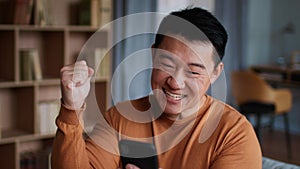Great news concept. Emotional middle aged asian man web surfing on smartphone and enjoying win, laughing at home