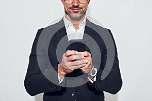Great news. Portrait of handsome and cheerful bearded man in classic wear and eyeglasses holding smart phone and looking