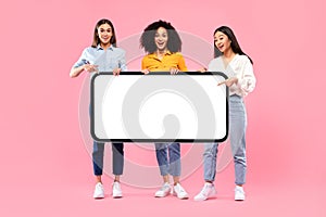 Great offer. Happy multiracial ladies friends holding and pointing at huge blank phone screen, pink background, mockup