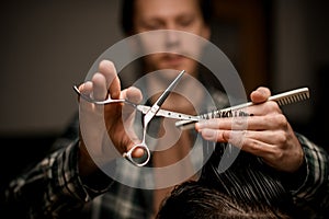 great selective focus on scissors in the hands of barber who cuts hair of client in barbershop.