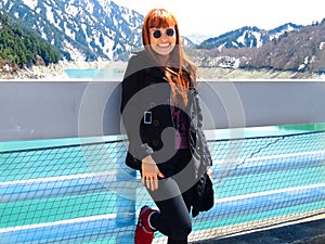 Pretty redhead woman smiling leaning agaisnt a bridge over a wonderful mountain background during a tour in Tateyama Kurobe Alpine