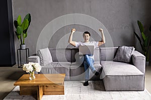 Great thing. Euphoric young man sit on couch by laptop screen shout super raise fists in winner gesture get good news job offer