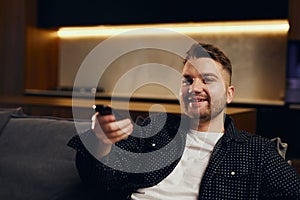 Great TV show. Handsome cheerful young man holding remote control and watching TV while sitting on sofa at home