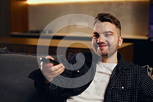 Great TV show. Handsome cheerful young man holding remote control and watching TV while sitting on sofa at home