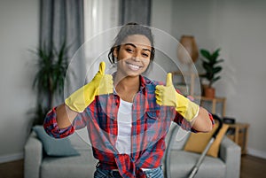 Great work. Happy young black lady in gloves shows thumbs up, enjoys cleaning result in living room interior