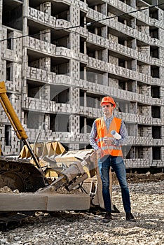 a great worker in uniform and helmet stands by an excavator on a construction site.