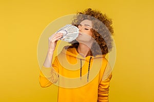 Greed for money. Portrait of avaricious curly-haired woman in urban style hoodie smelling dollar banknotes