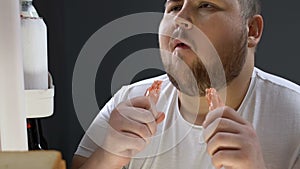 Greedy obese man eating sausages plastic cover near fridge at night, consumerism