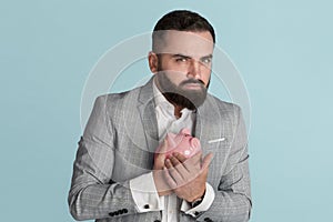 Greedy young businessman hugging piggy bank against blue background