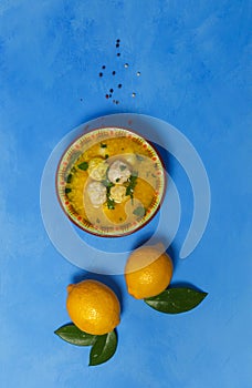 Greek avgolemono soup in a bowl and two lemons with leaves, on a bright blue background. Top view, copy of the space, vertically