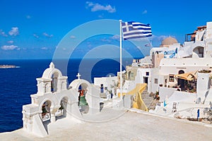 Greek orthodox church with bells and greek flag against famous white houses on Santorini island, Aegean sea, Greece