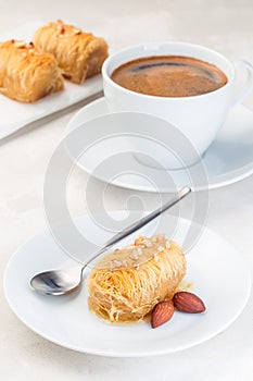 Greek pastry Kataifi with shredded filo dough stuffed with almond nuts, in honey syrup, on a white plate, served with cup of