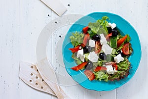 Greek salad with fresh vegetables, feta cheese and black olives in a blue plate on white wooden table.
